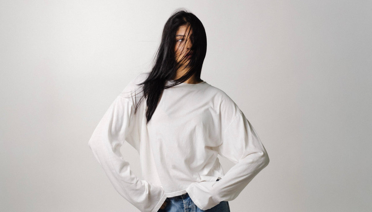 A woman with long black hair wearing an oversized white long-sleeve shirt and blue jeans, standing against a plain light gray studio background.