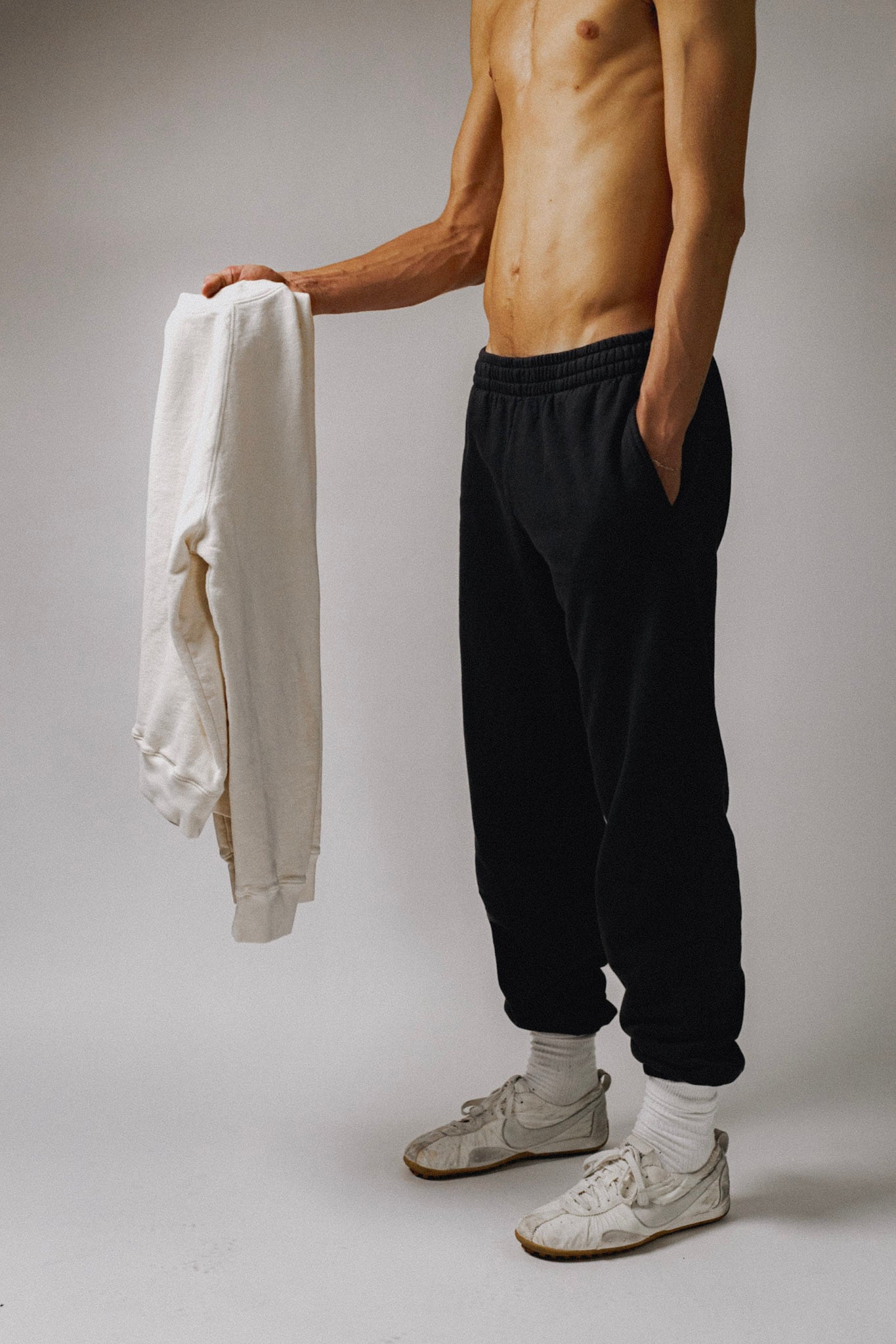 A shirtless model holds the Legacy Raglan in Bone and wears the Legacy Sweatpant in True Black in front of a white seamless backdrop wearing white sneakers.