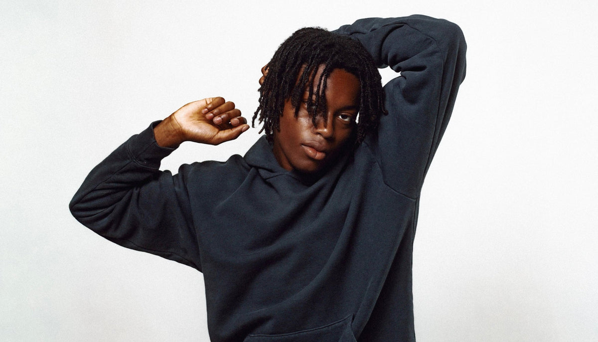 A person with dark dreadlocks wearing a black hoodie, posing with one arm raised against a plain light gray background.